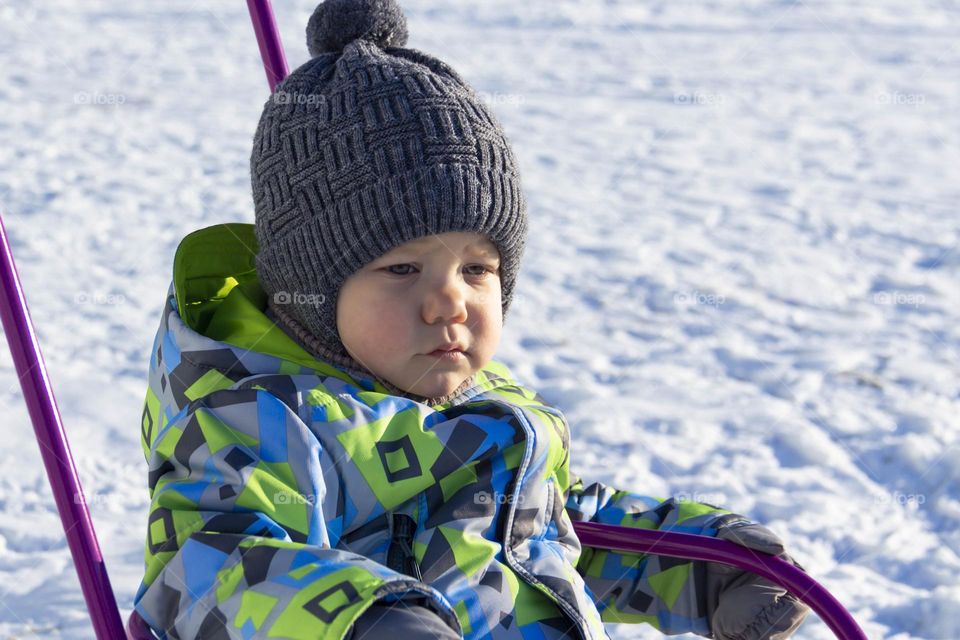A child in winter in winter jackets, pants, a hat and boots on white snow on the street and in the park in nature plays winter fun and sleds.