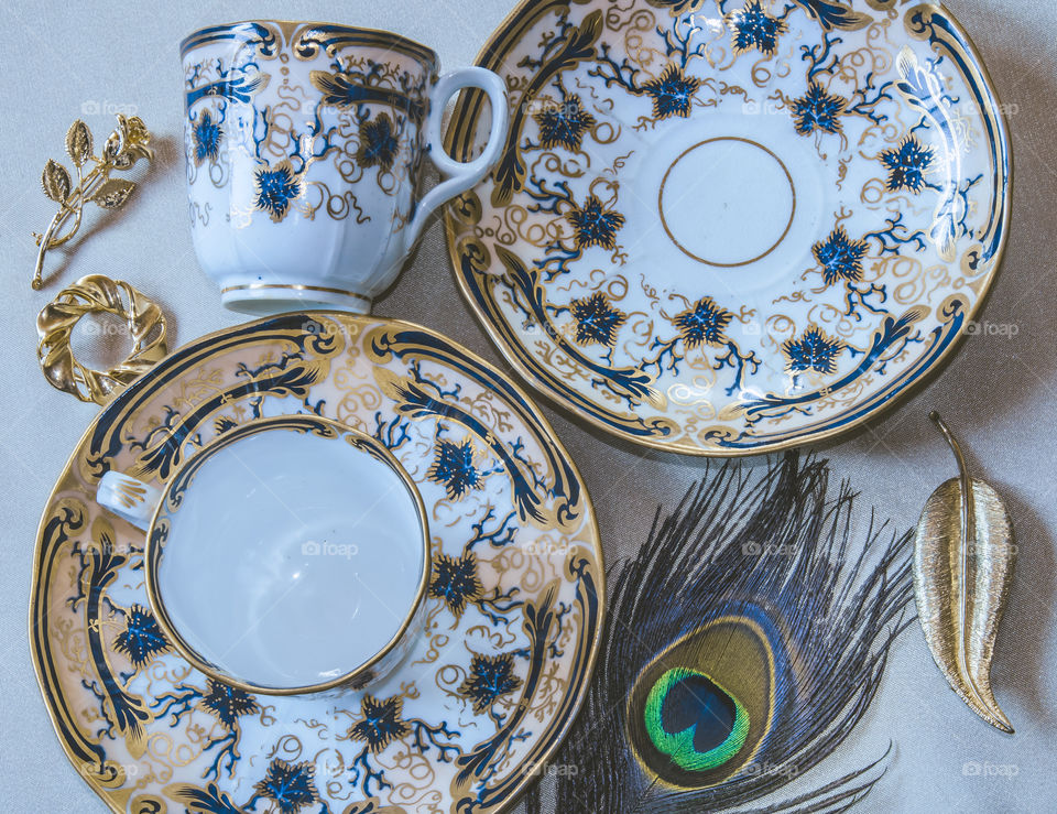 Flat lay featuring antique Davenport blue and gold teacups and saucers, gold brooches and a peacock feather