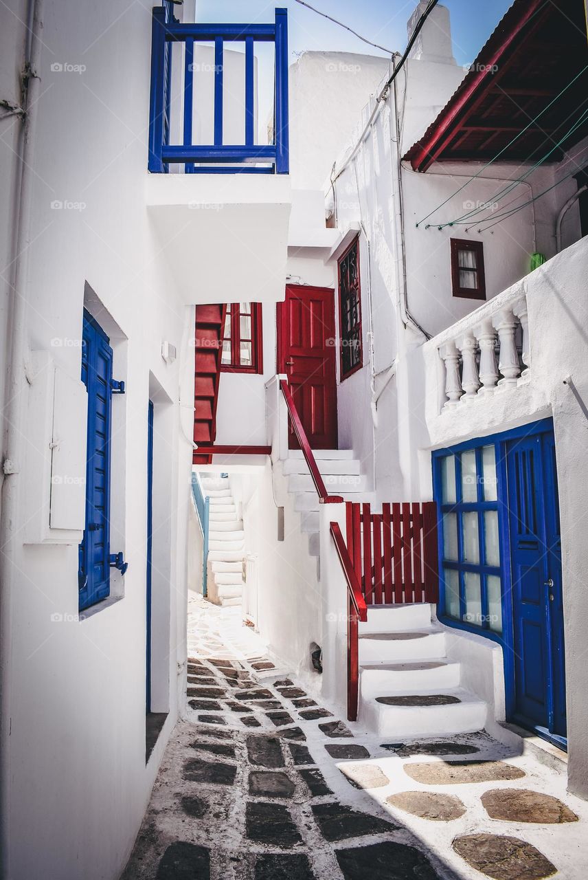 The bright white buildings lighten the narrow streets of Mykonos, Greece. Bright blue and red accents bring the scene to life.