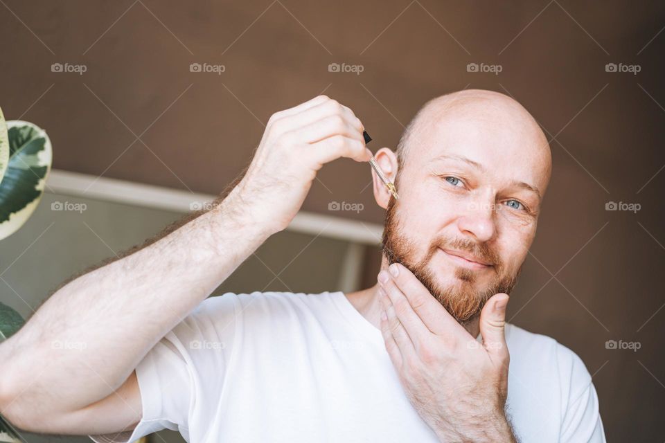 Adult handsome man with pipette with beard oil in bathroom at home