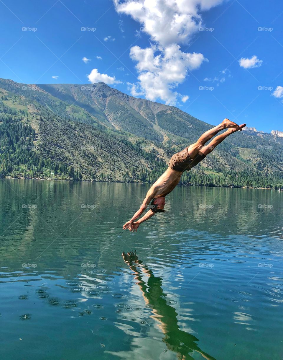 Young millennial jumps off rope swing and dives into the beautiful clear blue lake with mountains reflecting off water on summer vacation 