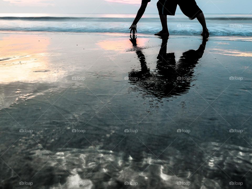 Reflection of a man picking sea shells on a wet sandy beach in the evening