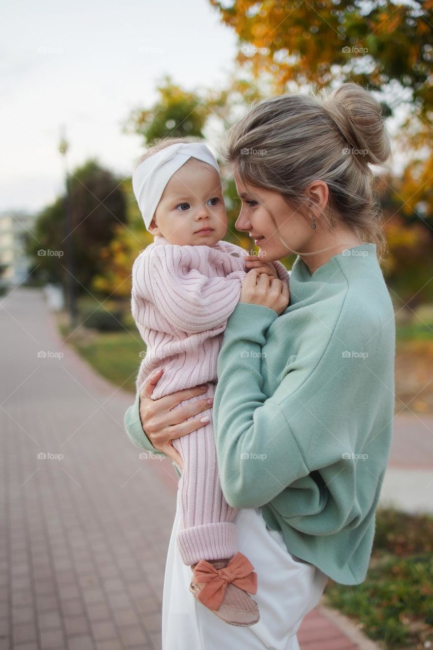 Young stylish mother holds the child in her arms, hugs in an autumn park. Mother in a mint jacket and a baby girl mint knitted suit and a headband. Concept: motherhood, mothers day. Baby 10 months