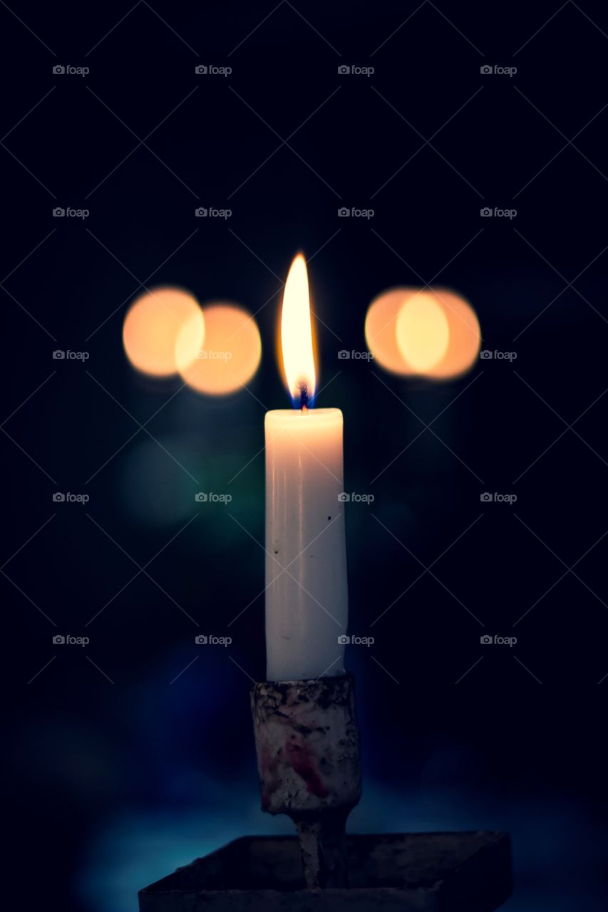 A moody portrait of a single candle surrounded by darkness in the evening with in the background some bokeh balls of other candles.