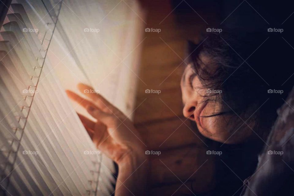 Portrait of young woman looking through window blinds. The person seemed curious about something outside. The window curtains are white and have thin, horizontal slats in low angle view