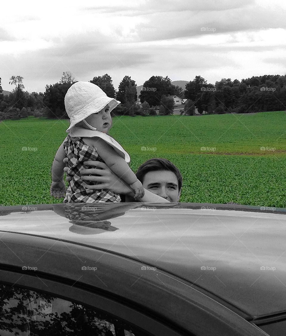 Hey, what am I doing up here?  Baby girl sitting atop a car with a scenic green field behind her.  Photo taken in rural Vermont.  
