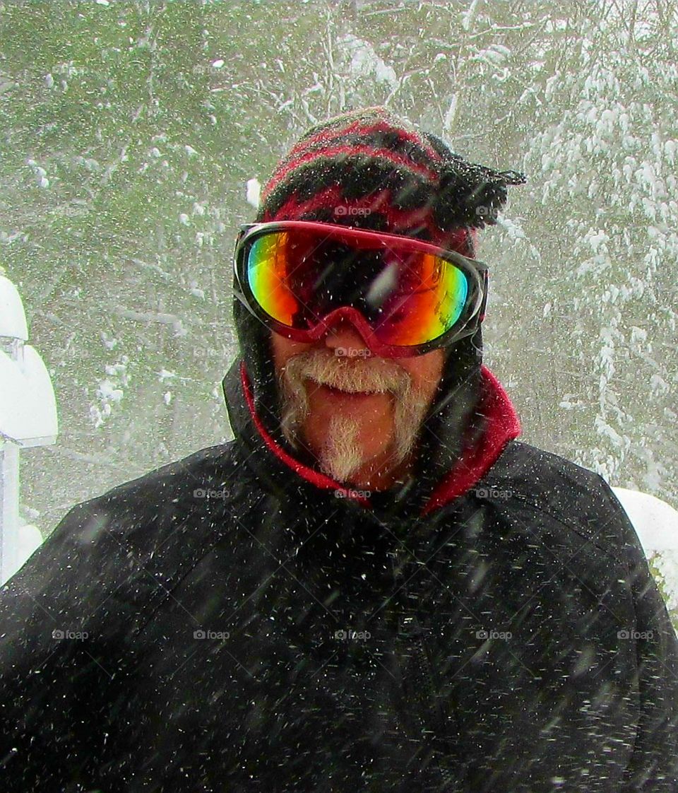 Man in snow wearing ski goggles, ski wear, smiling, moustache, as it's snowing out.