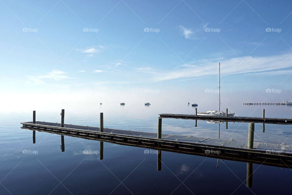 Sailboat reflection on calm water with blue sky with some clouds and white fog