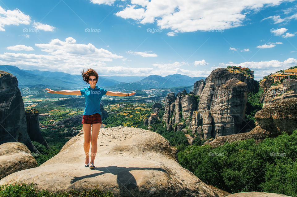 Young happy woman stands back to the rock with raised hands. Traveler enjoying the landscape with mountain. Monasteries of Meteora, Greece
