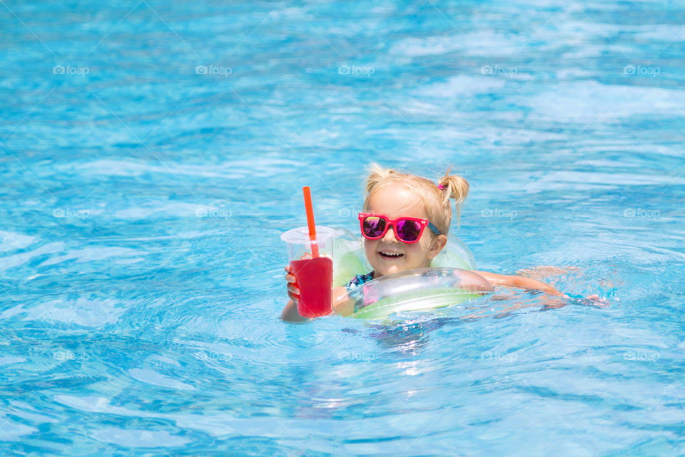 Little girl with blonde hair in swimming pool with fresh juice 