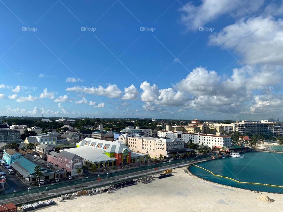 A landscape view of a town at port aboard a cruise ship. Blue skies with white clouds and a sandy beach invite tourists. 