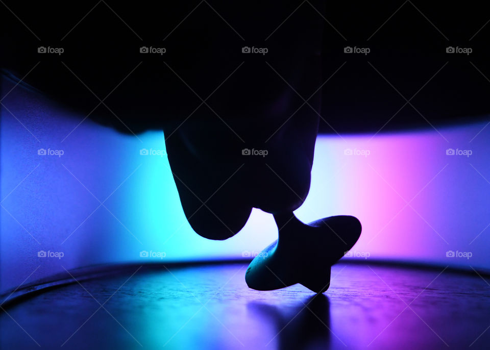 A silhouette of a hand holding a metal top preparing to spin it on a coffee table in a psychedelic cosmic colorful neon bright enclosing.