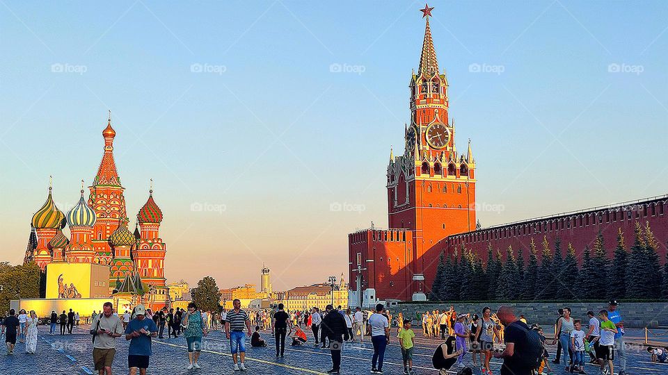 Summer evening.  Red Square where people walk.  Basil's Cathedral and the Spasskaya Tower with chimes in the orange rays of the setting sun. Moscow