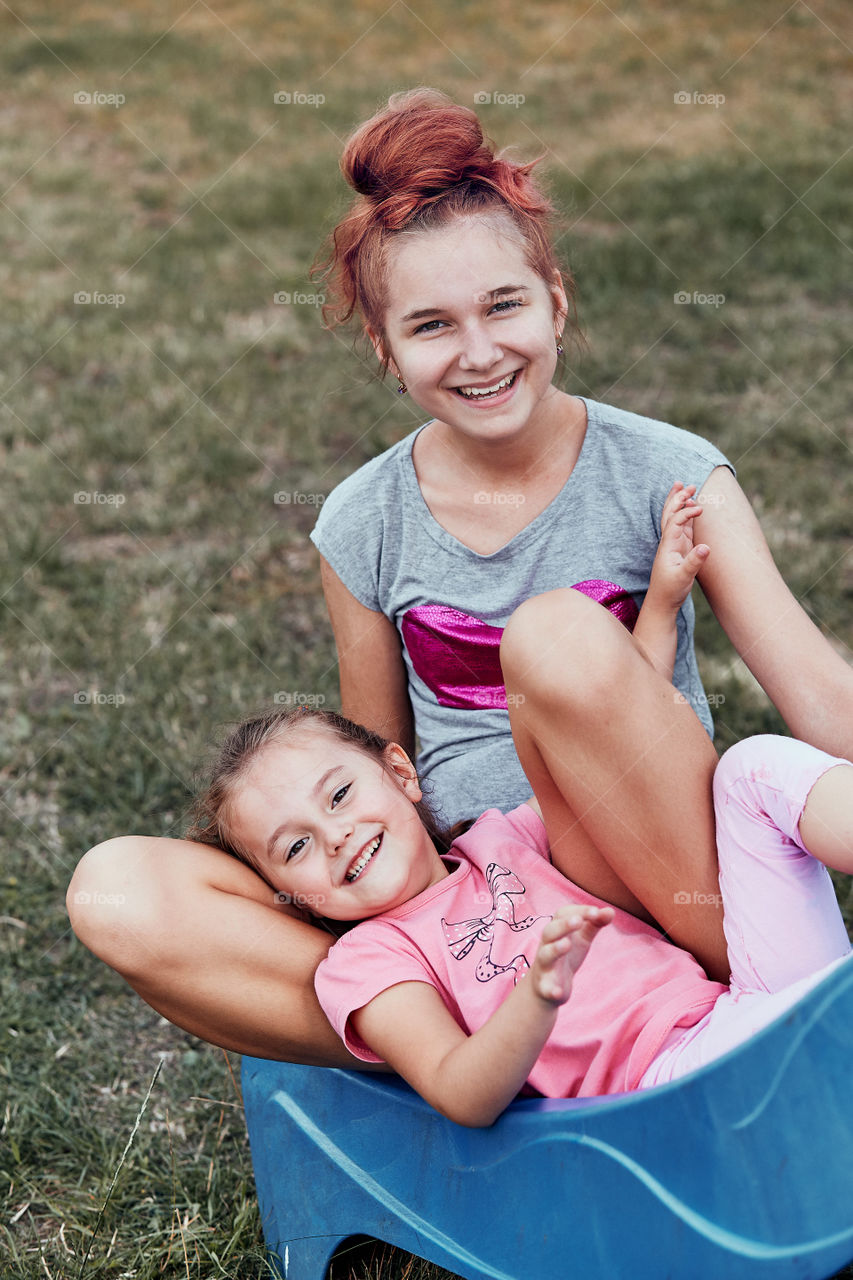 Teenage girl playing with her younger sister in a home playground in a backyard. Happy smiling sisters having fun on a slide together on summer day. Real people, authentic situations