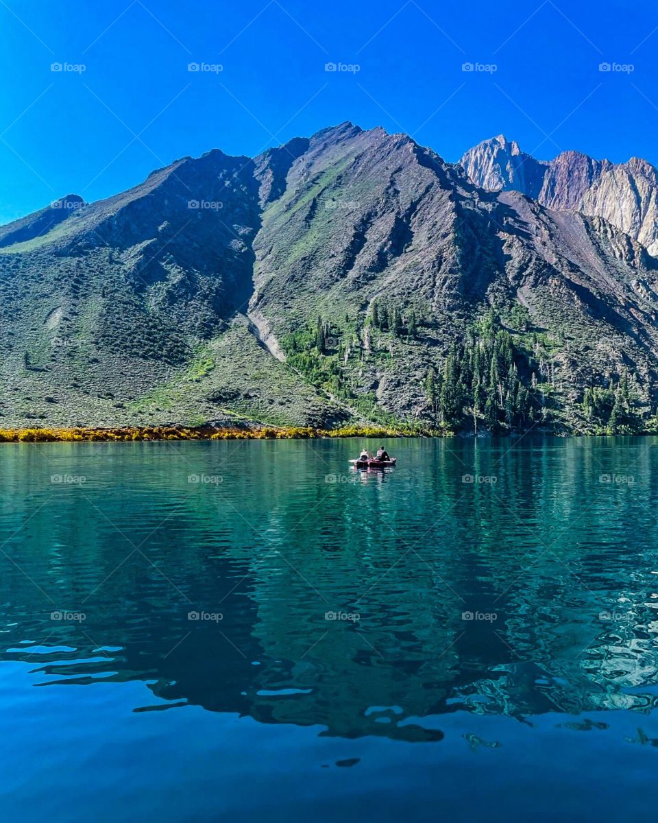 Gray rock mountains above a blue lake with a touch of yellow foilage, with two men in a boat. 