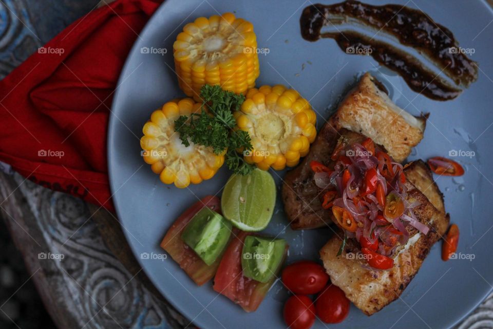 Tuna steak cooked with corn garnished with cherries, sliced tomatoes and lemon topped with sliced hot chili peppers on a baby blue melamine plate lined with red cloth and thick wood. Restaurant food.