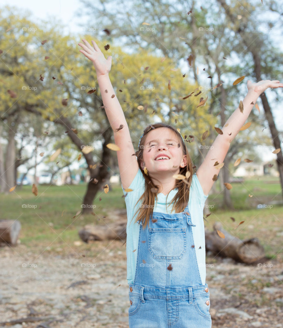 Girl in overalls tossing small leaves with arms in the air.