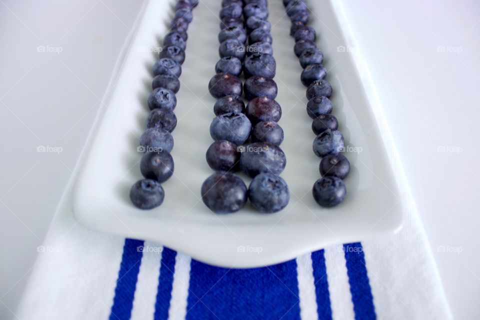 Angled view of blueberries on a rectangular white plate, arranged in lines to emphasize the blue stripes on the white dish towel underneath, all on a white background 
