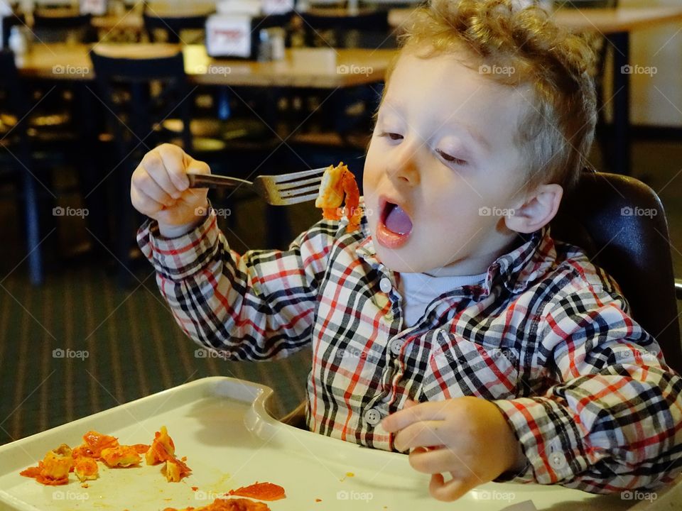 A little boy sitting in a high chair digs into his favorite snack of pepperoni pizza with great vigor. 