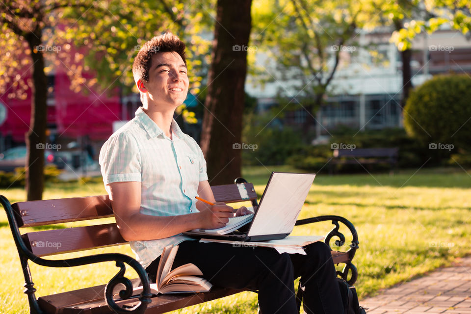 Student working on a laptop using books and notes sitting on a bench in a park. Young boy wearing a blue shirt and dark jeans