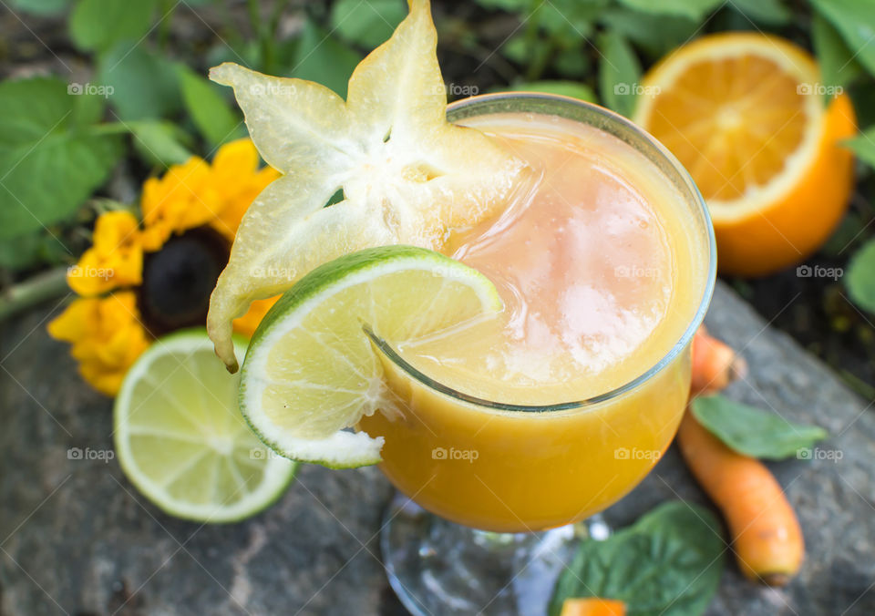 High angle view of mixed flavor fruit and vegetable creamy tropical smoothie with pink berry smoothie center and colorful smoothie healthy ingredients in background: carrots, lime, orange, sunflower, spinach, and tangy carambola star fruit 