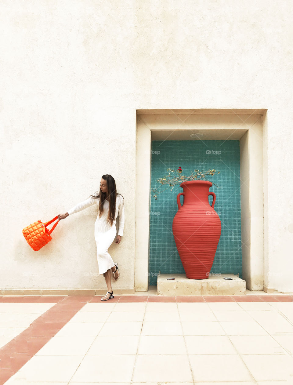 Young woman with long hair and fashionable accessories standing in front of a monochrome wall with a huge orange jar 