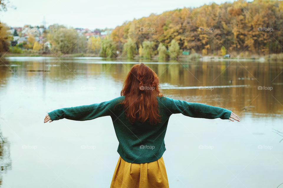 Beautiful portrait of young redhead curly woman standing near big lake in colorful autumn park in October. Fall season. Feeling freedom.