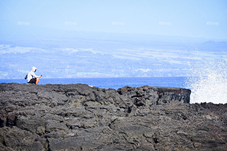 Watching the waves and using technology from the lava at Richardson Ocean Park in Hilo, Hawaii with the volcano rising up in the background.