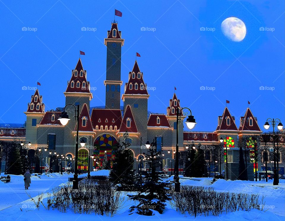 Winter. Amusement complex "Dream Island" made of stone, with towers, decorated with a luminous garland. Twilight. Above the complex is a large moon. In the foreground trees and firs in the snow