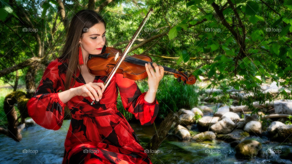 beautiful woman in red dress playing violin at the edge of a rocky river