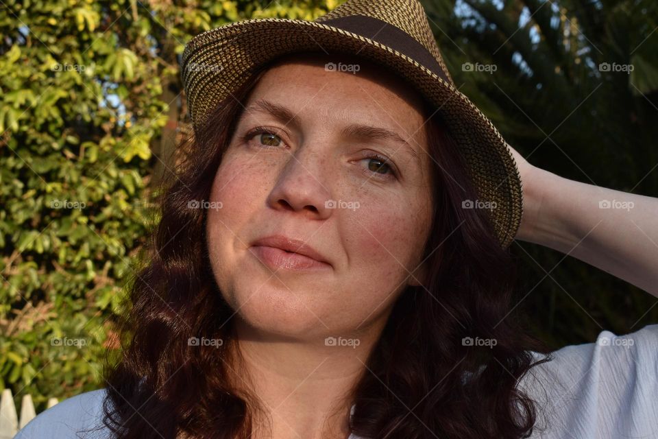 Women with natural and minimal makeup standing outdoors outside with wet hair. Women with red hair and green eyes standing next to tree taking a selfie. Sunlight shine on women hair and skin and making shadows. Women wearing a hat.