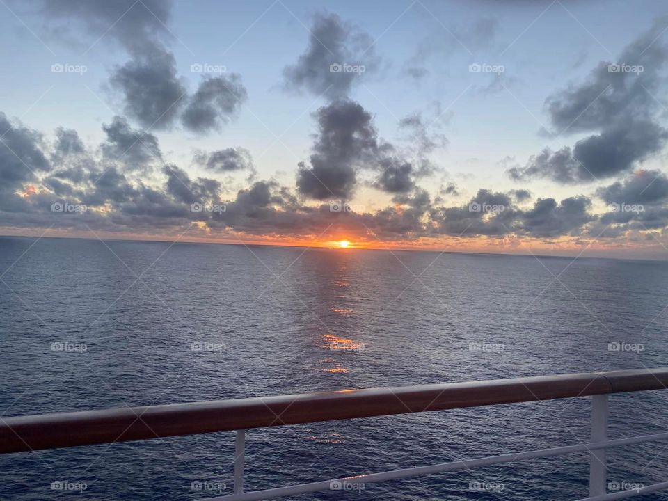 The orange sun peeks behind the silver blue waters of the ocean as it sets in the distance. The clouds are grey blue I. A light blue sky. A relaxing evening aboard a cruise ship. 