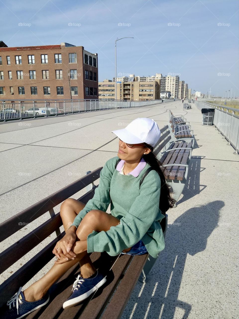 A woman sitting on the bench wearing a white baseball hat against the blue sky and buildings.