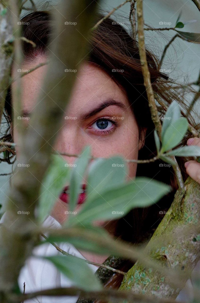 Young woman with brown hair, blue eyes, and dark red lipstick looking through branches and leaves.