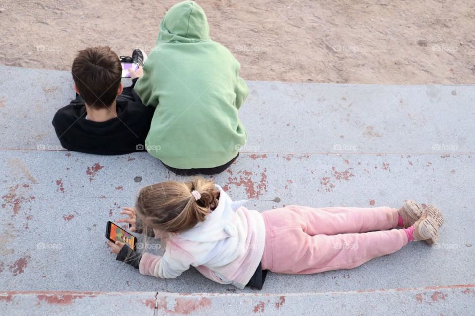 Three kids sitting on big stairs outdoor playing video games in smartphones