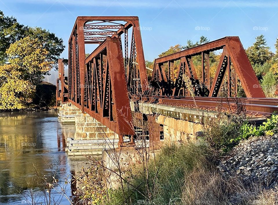 Rusty Iron Of A Train Trestle Spans A River And Contrasts With The bLue Sky.