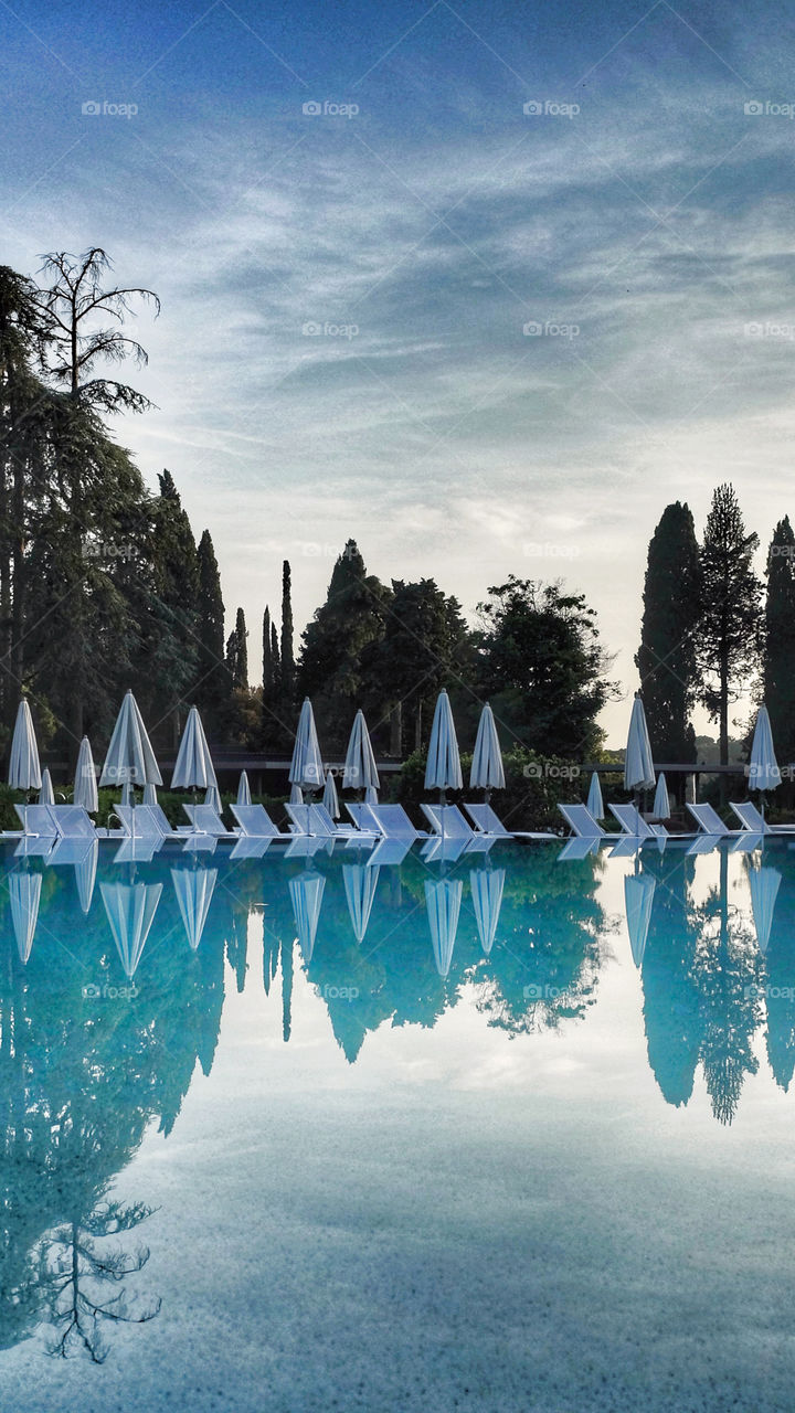 Reflections of trees, sun beds and parasols in a summer swimming pool at dusk.