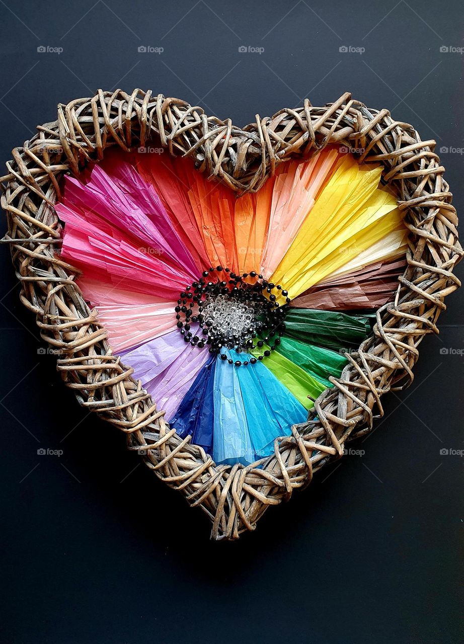 Flatlay composition of a heart-shaped flower made of multi-coloured silk papers, black and clear beads and a heart-shaped wicker wreath, placed on black paper and creating a colour explosion