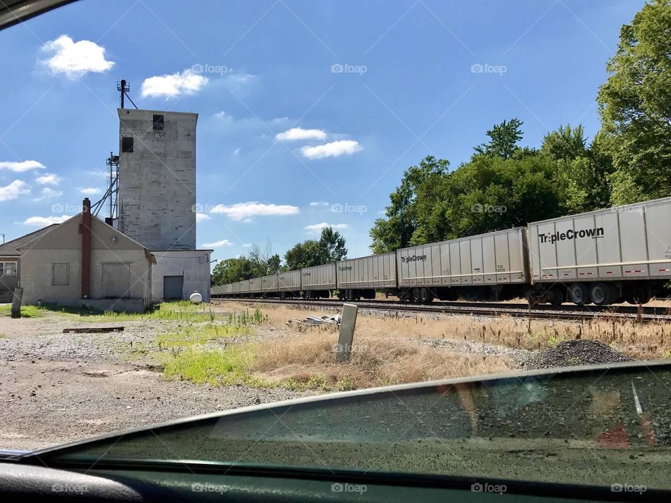 A train passes a white building with a blue sky, white clouds, and green trees in the background. 