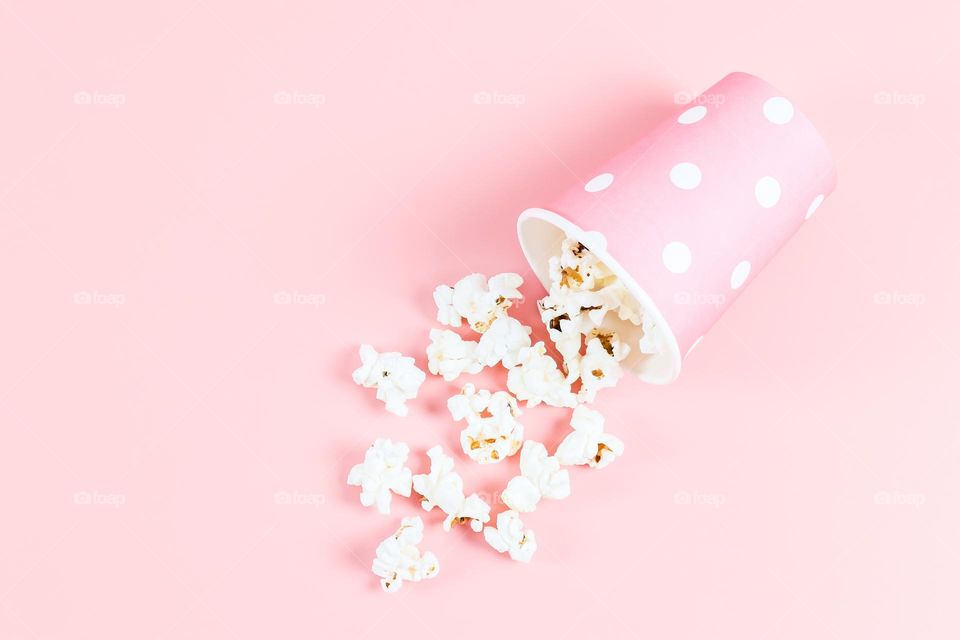 One pink paper glass with white polka dots with popcorn spilling out of it lies on a soft pink background, flat lay close-up.
Оставить