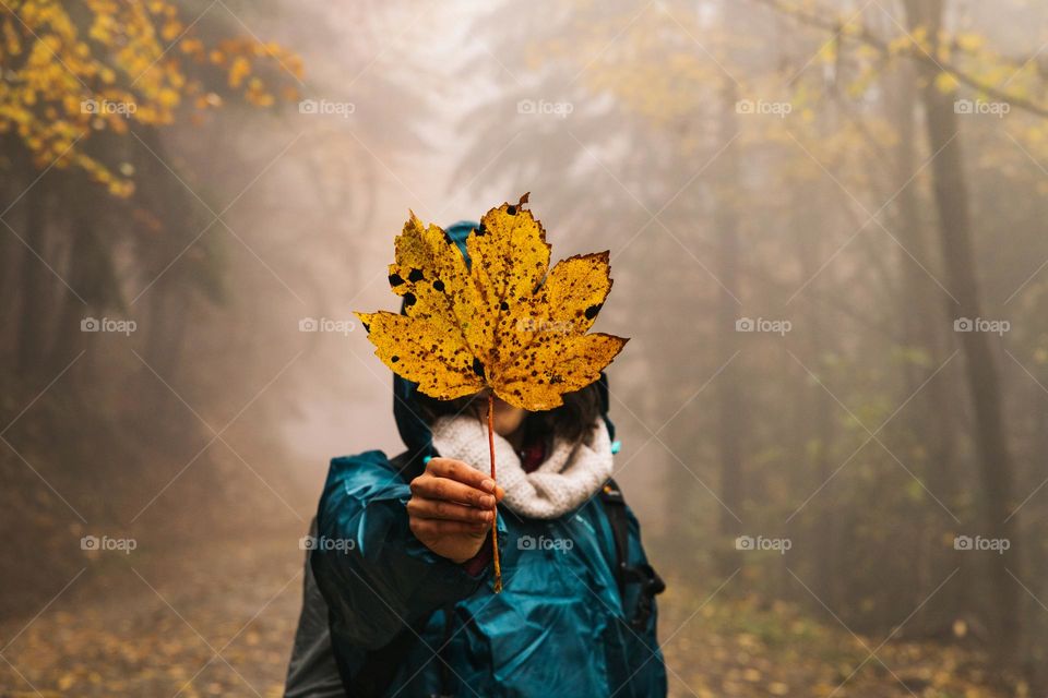 Woman holding a big yellow leaf, while being in nature, hiking, on a autumn rainy day.