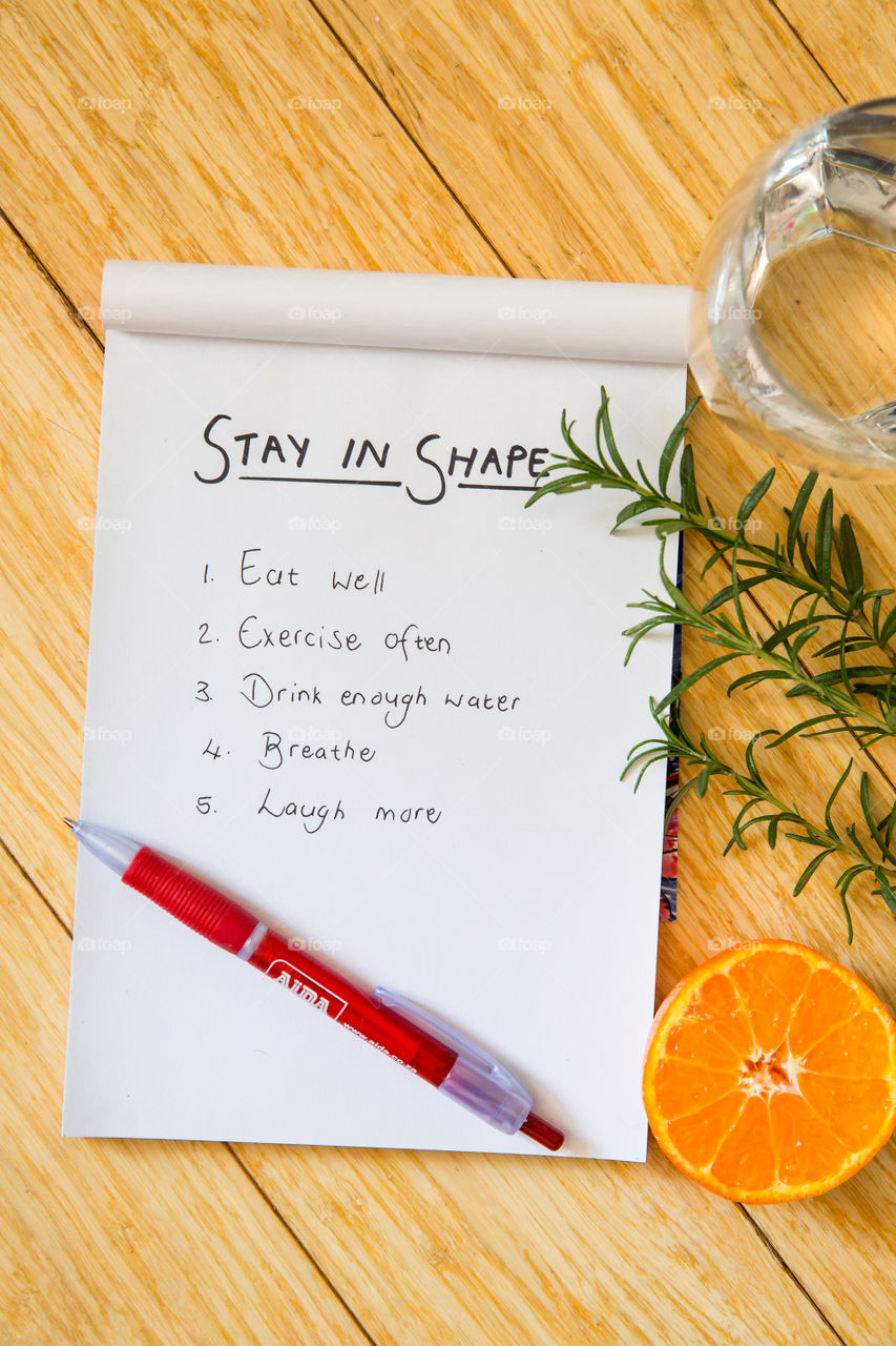 Handwritten list on notepad to encourage one to stay in good shape. Things to do to stay on track to be healthy physically and mentally. Lay flat with water, fruit, herbs and a notepad on wooden background.