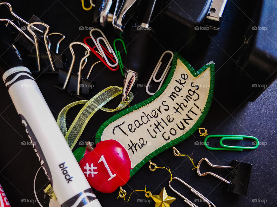 Teacher Appreciation. Teachers make the little things count flatlay on a black background surrounded by teachers school supplies including paper clips, a black stapler, a hole puncher, black binder clips, a black pen and a black marker.