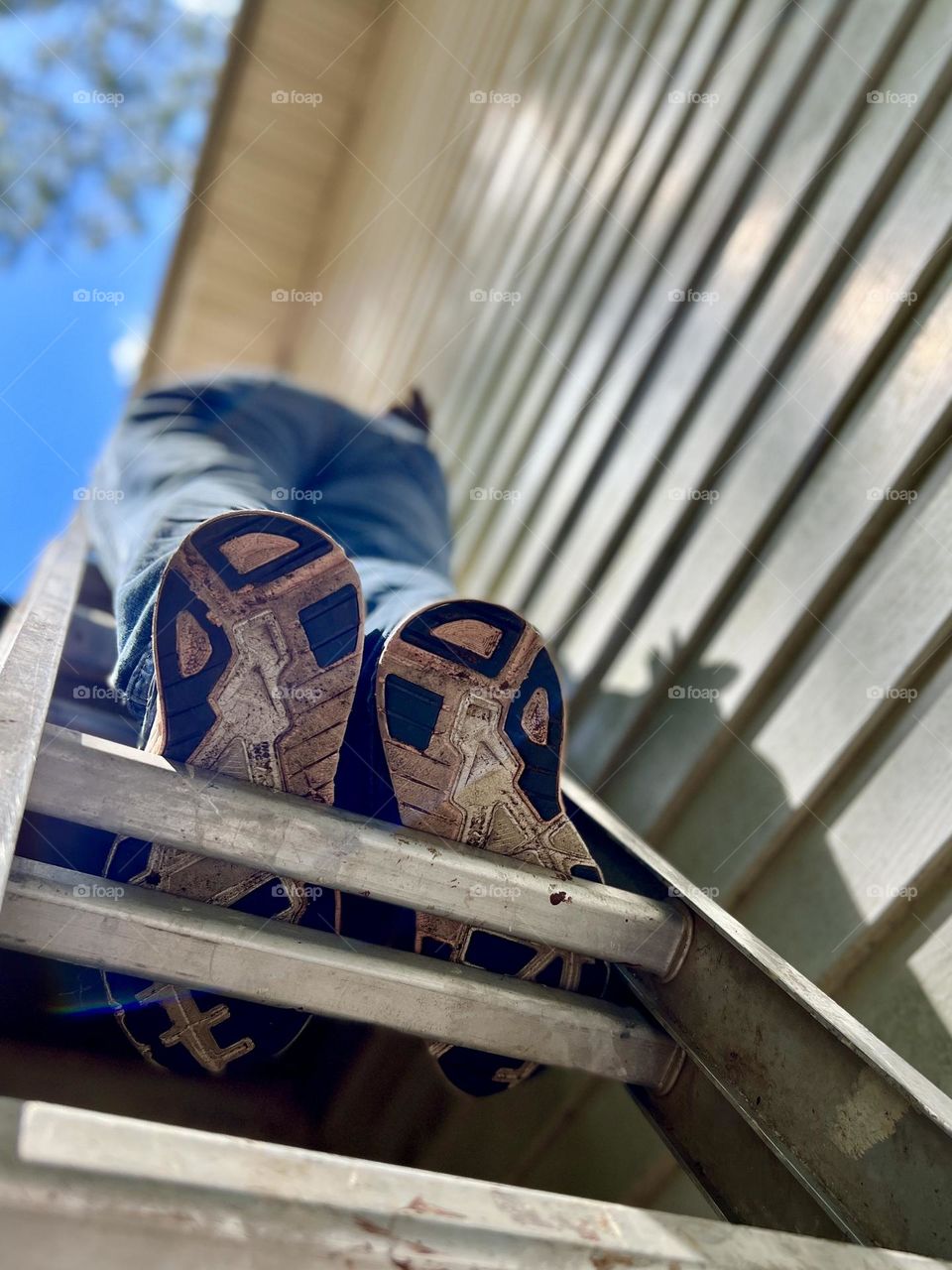 Looking up at person on a ladder. The sky, siding on the building and person’s shadow.