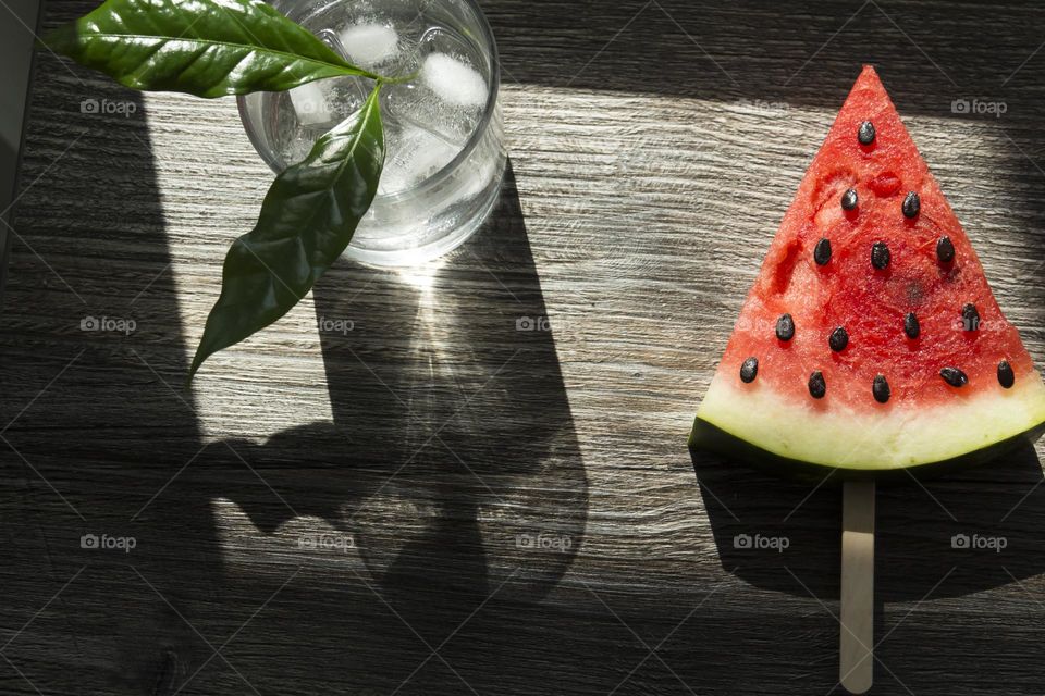 A piece of ripe, red watermelon and a cold cocktail with ice and green leaves are on a wooden table.  Summer drinks and snacks.