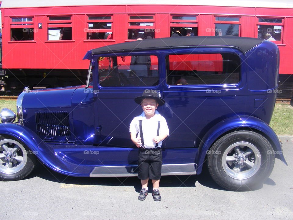 Young and Old. An excited young  boy stands proudly in front of an old classic blue motor vehicle. Behind is a red carriage of an old steam train. It is Art Deco weekend in Napier NZ
