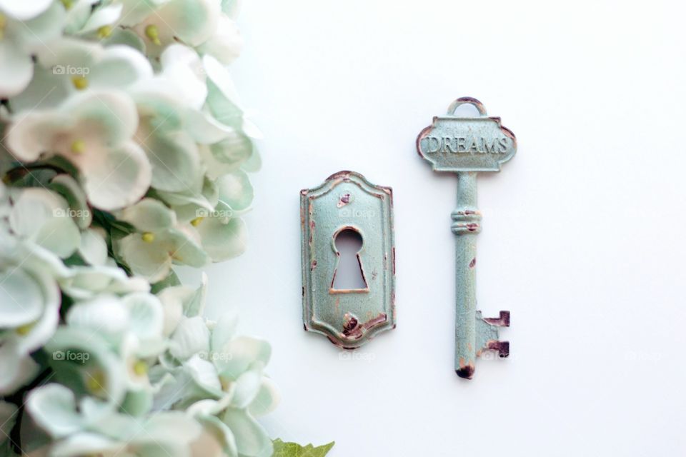 Overhead view of light teal, vintage, metal key with embossed word, “Dreams,” and keyhole with light teal hydrangeas on white background - landscape