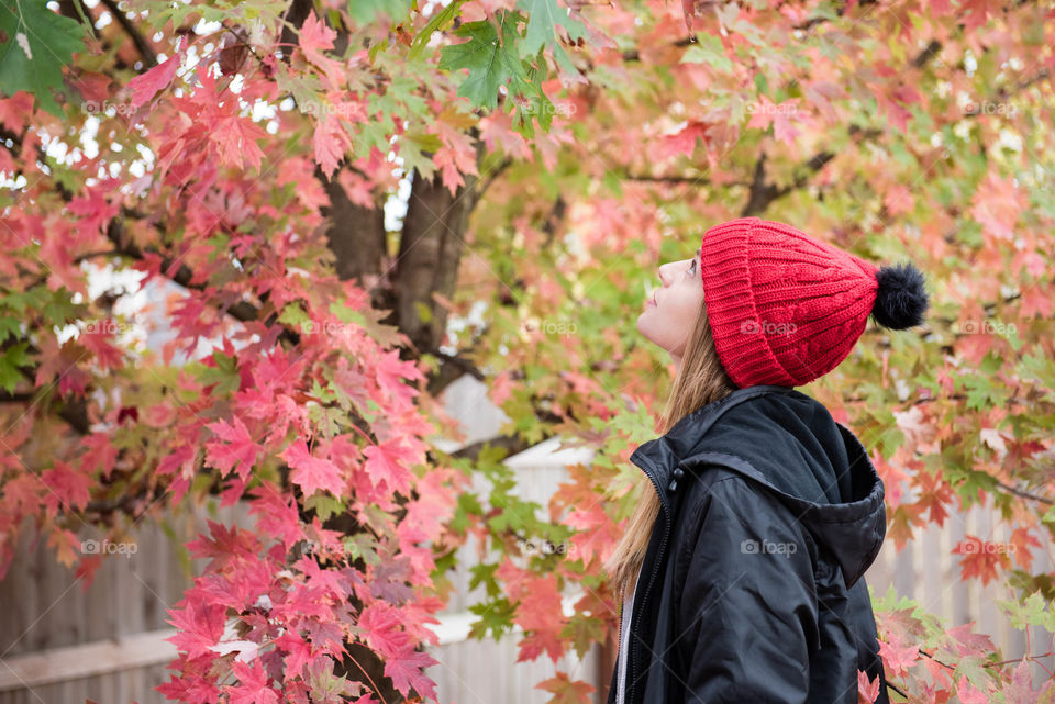 Young millennial woman wearing a pom pom knit hat and coat and looking up at a tree with fall leaves