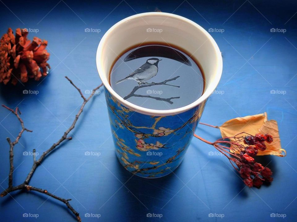 On a dark blue background with a reflective effect, there is a mug with a Japanese-style pattern. In a mug of black coffee on the surface of which is a reflection of a cherry branch and a bird. Next to the mug lies a cherry branch and a branch with