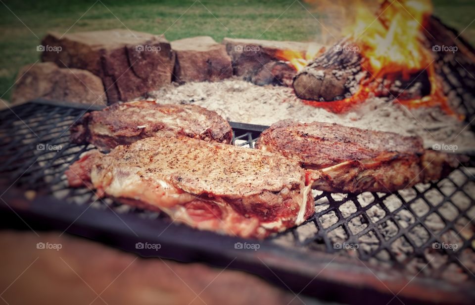 Steaks cooking outside in an open fire pit over hot coals with a fire burning in the background
